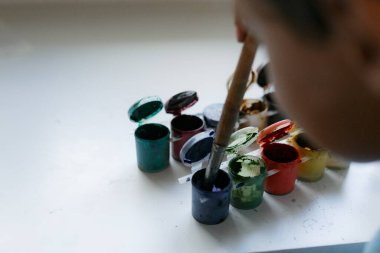 Mavi tişörtlü kıvırcık saçlı çocuk beyaz bir masada otururken boyalı bir resim çiziyor.