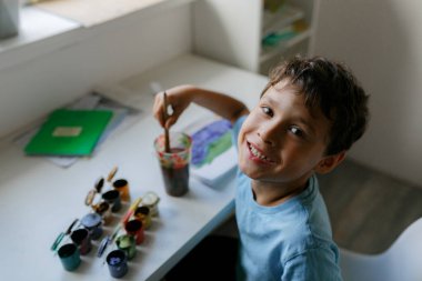 Mavi tişörtlü kıvırcık saçlı çocuk beyaz bir masada otururken boyalı bir resim çiziyor.
