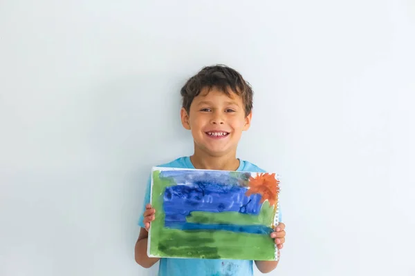 Mavi tişörtlü kıvırcık saçlı çocuk beyaz bir masada otururken boyalı bir resim çiziyor.