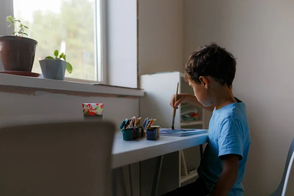 Mavi tişörtlü kıvırcık saçlı çocuk beyaz bir masada otururken boyalı bir resim çiziyor.