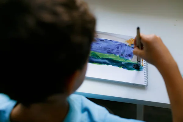 Mavi tişörtlü kıvırcık saçlı çocuk beyaz bir masada otururken boyalı bir resim çiziyor.