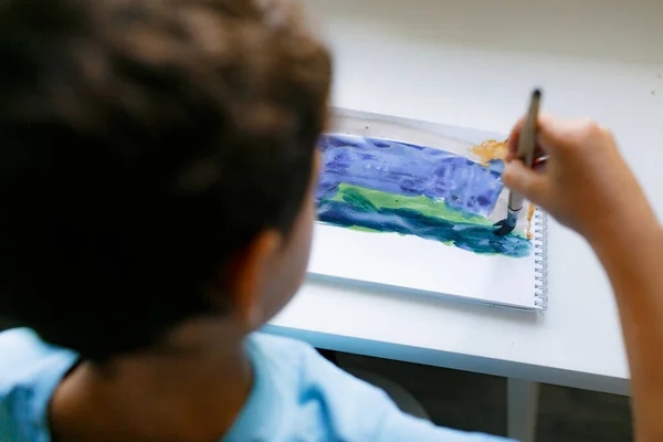 Mavi tişörtlü kıvırcık saçlı çocuk beyaz bir masada otururken boyalı bir resim çiziyor.