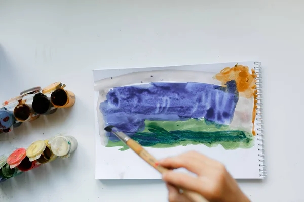 Mavi tişörtlü kıvırcık saçlı çocuk beyaz bir masada otururken boyalı bir resim çiziyor.