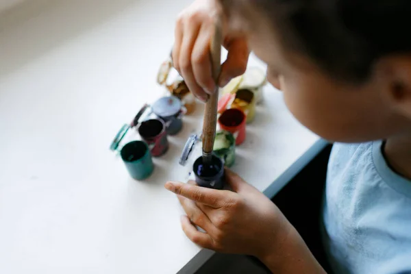 Mavi tişörtlü kıvırcık saçlı çocuk beyaz bir masada otururken boyalı bir resim çiziyor.