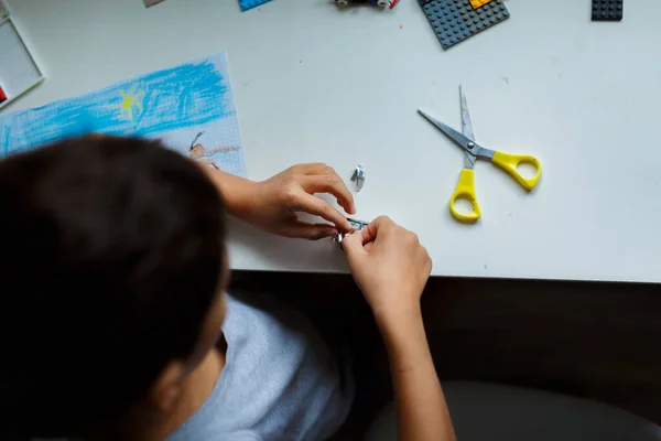 Gri tişörtlü, elinde makasla oturan Asyalı siyah saçlı çocuk farklı şekillerde kesilmiş.