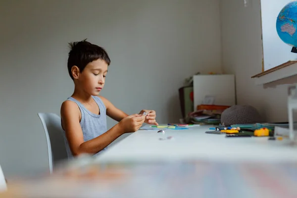 Koyu renk saçlı çocuk beyaz bir masada otururken kalemlerle resim çiziyor.