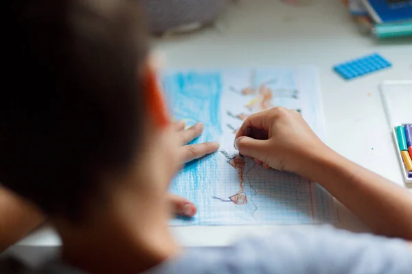 Koyu renk saçlı çocuk beyaz bir masada otururken kalemlerle resim çiziyor.
