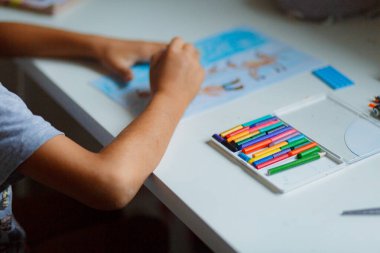 Koyu renk saçlı çocuk beyaz bir masada otururken kalemlerle resim çiziyor.