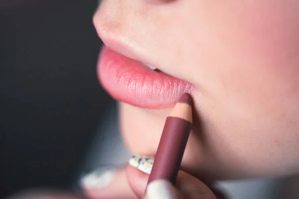 Woman putting on makeup, painting lips with red lipliner. Closeup of lips touched with pencil, studio. Small depth of field.