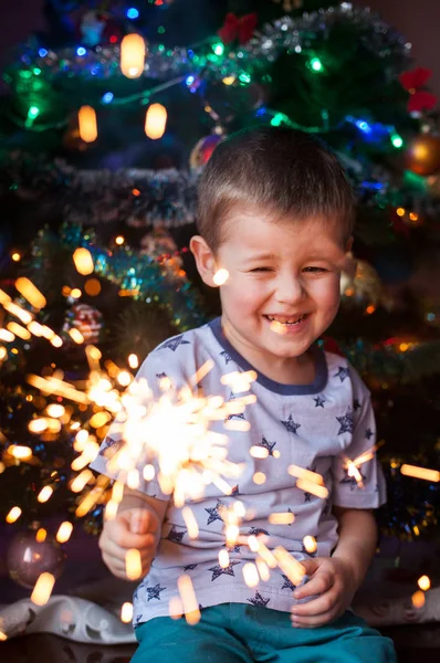 Schönes kleines Kind mit brennender Wunderkerze am Silvesterabend, Bengalfeuer. es gibt einen Hintergrund mit bunten Lichtern. — Stockfoto