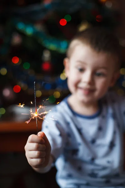 Schönes kleines Kind mit brennender Wunderkerze am Silvesterabend, Bengalfeuer. Der Schwerpunkt liegt auf Feuerwerk. glücklicher Junge mit Wunderkerzen auf verschwommenem Weihnachtshintergrund. — Stockfoto