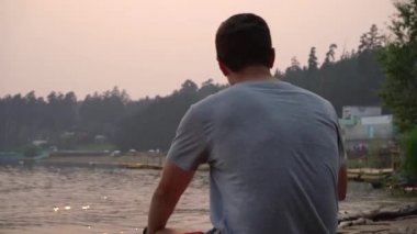 Genç bir adam gün batımı ve kıyı şeridine karşı oturuyor..