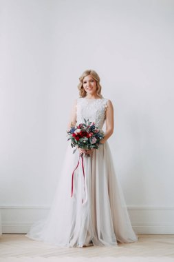 Parlak bir stüdyoda gelinlik içinde güzel bir gelin. Avrupa usulü düğün. Dekor ve buket elinde. Genç kız ayakta, oturuyor ve poz veriyor.