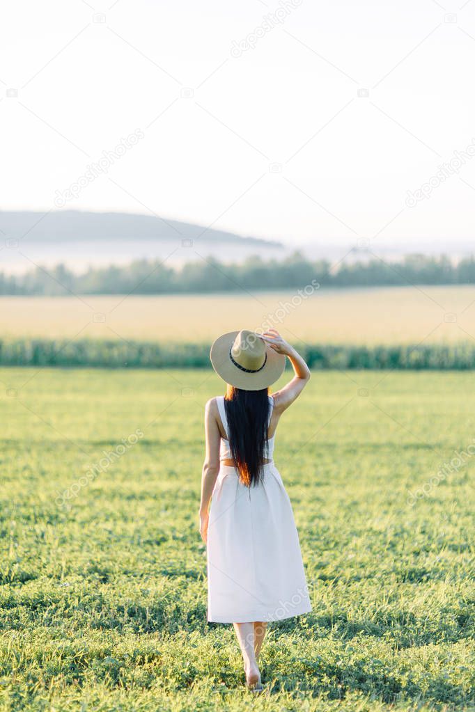 Girl walking on the field, in a hat and summer dress. Smiling and laughing, beautiful sunset in the forest and in nature. White dress and rye, sloping fields. Happy traveler, lifestyle.