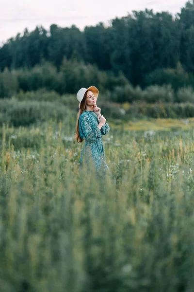 帽子と夏に美しいロシアの女の子のドレス 腰に草のフィールド上を歩いてします 幸せな旅行者 収集するハーブ 笑みを浮かべてします 森の夕日 — ストック写真