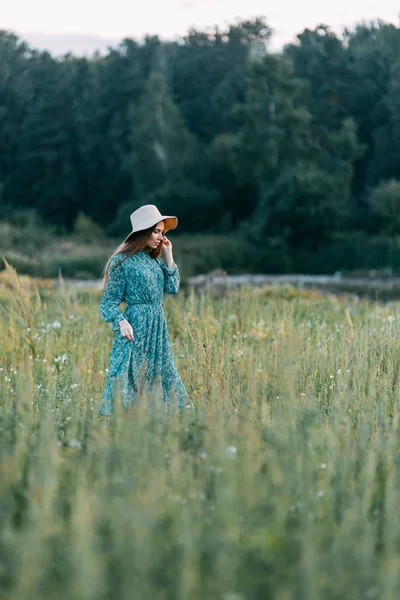 帽子と夏に美しいロシアの女の子のドレス 腰に草のフィールド上を歩いてします 幸せな旅行者 収集するハーブ 笑みを浮かべてします 森の夕日 — ストック写真