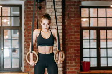 Sportif kız gücü eğitim uygulamaları için eğitim salonunda. Yüzük, ağırlık ve dumbell elinde. Gülümseyen ve spor oynarken.