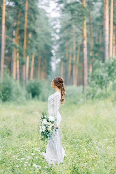 The bride in a wedding dress in nature with a bouquet. Boudoir photo shoot in pine lemu in the style of fine art.