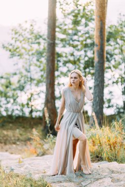Bir çam ormanı içinde boudoir elbiseli kız. İnce bir uçan elbise kayalarda tutuyoruz. Düğünde doğa ile birlik. Gün batımında gelin