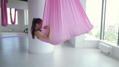 Genç bir atletik kız hamakta yoga yapıyor, sağlık egzersizleri yapıyor. Spor salonunda panoramik pencerelerle çalışma.