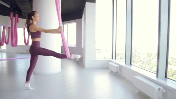 Una giovane ragazza atletica fa yoga su un'amaca, esercizi per la salute. Allenamento in palestra con finestre panoramiche. — Video Stock