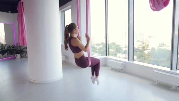 En ung atletisk flicka gör yoga på en hängmatta, övningar för hälsan. Träning i gymmet med panorama Windows. — Stockvideo