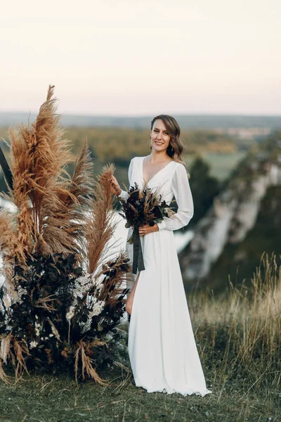 La novia con un ramo de flores se encuentra en un arco triangular. Ceremonia de boda en estilo boho. —  Fotos de Stock