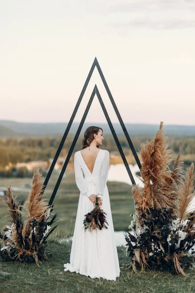 La novia con un ramo de flores se encuentra en un arco triangular. Ceremonia de boda en estilo boho. —  Fotos de Stock