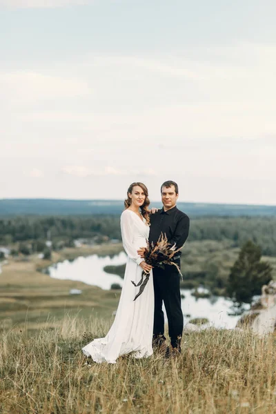 Egy gyönyörű tündérmese boldog menyasszony egy csokorral, koszorúval és egy elegáns vőlegény a háttér sziklák a hegyekben. Boho esküvő a természet. — Stock Fotó