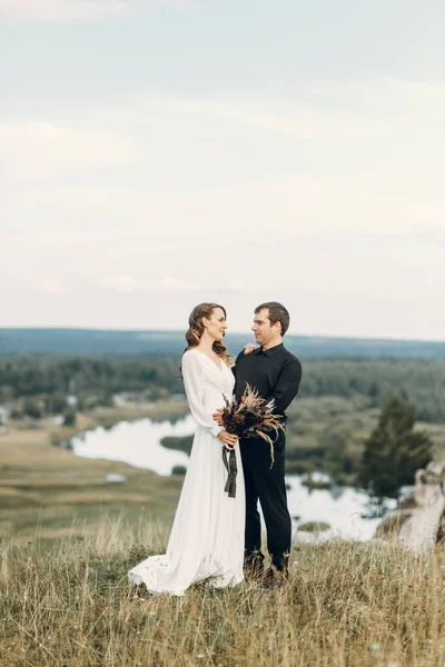 Una bella sposa felice fiaba con un bouquet e una ghirlanda e uno sposo elegante sullo sfondo di rocce in montagna. Boho matrimonio in natura. — Foto Stock