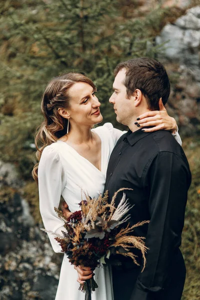 Uma linda noiva feliz de conto de fadas com um buquê e uma grinalda e um noivo elegante no fundo de rochas nas montanhas. Boho casamento na natureza. — Fotografia de Stock