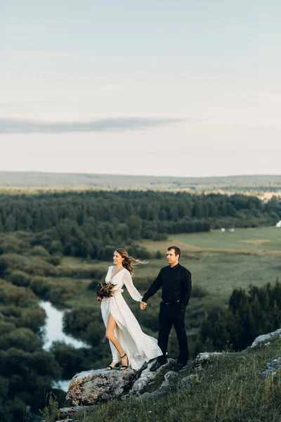 Uma linda noiva feliz de conto de fadas com um buquê e uma grinalda e um noivo elegante no fundo de rochas nas montanhas. Boho casamento na natureza. — Fotografia de Stock