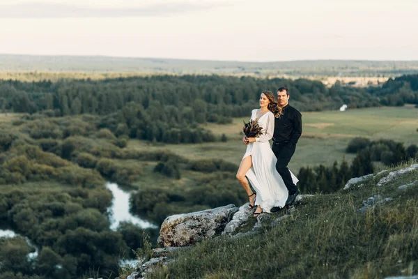 Uma linda noiva feliz de conto de fadas com um buquê e uma grinalda e um noivo elegante no fundo de rochas nas montanhas. Boho casamento na natureza. — Fotografia de Stock