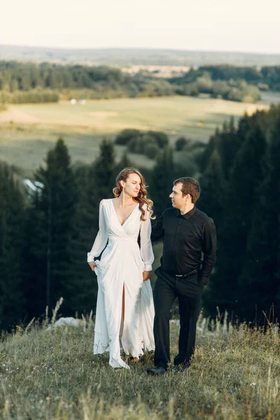 Uma linda noiva feliz de conto de fadas com um buquê e uma grinalda e um noivo elegante no fundo de rochas nas montanhas. Boho casamento na natureza. — Fotografia de Stock