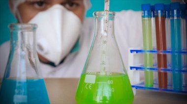 Simyacı, şişeler tutan, bir testin farklı kompozisyonlar, beyaz önlük, solunum, Gözlük, beyaz eldivenleri, yeşil bir arka plan ile test tüpleri testleri.