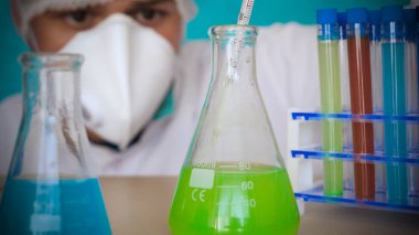 Simyacı, şişeler tutan, bir testin farklı kompozisyonlar, beyaz önlük, solunum, Gözlük, beyaz eldivenleri, yeşil bir arka plan ile test tüpleri testleri.