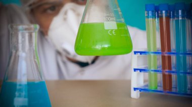 Simyacı, şişeler tutan, bir testin farklı kompozisyonlar, beyaz önlük, solunum, Gözlük, beyaz eldivenleri, yeşil bir arka plan ile test tüpleri testleri.