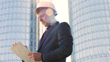 Başarılı genç yakışıklı bir mühendis, mimar, Oluşturucu, bir defter, gökdelen arka plan yazma, takım elbiseli, beyaz bir kask giyen işadamı portresi.