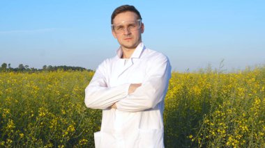 Genç yakışıklı biyolog veya beyaz tıbbi kıyafetler, koruyucu gözlük, kanola, gülümseyen, başarılı, alan ziraat mühendisi portresi biyo.