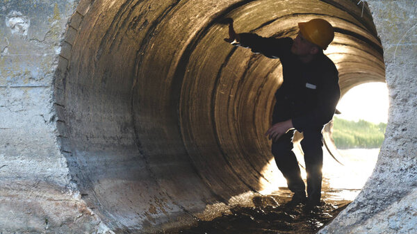 Engineer, worker checks gutters, in blue robe, in boots, in a construction yellow helmet, tunnels, technical supervision.