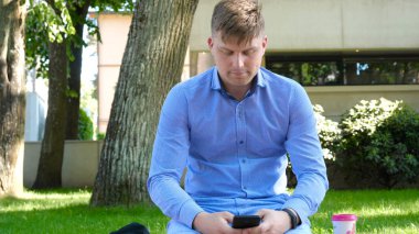 Parkta oturan genç yakışıklı öğrenci adam telefon ve içecekler kahve mesajlar yazıyor. Konsept: çalışma, dinlenme, iletişim, sohbet.