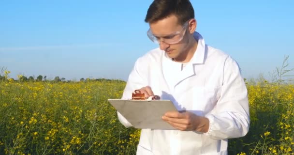 若いハンサムな 生物学者またはアグロノミストは 白いコート ゴーグル 青いゴム手袋で 水分の葉 Dna ピペットの分析を取り リンゴの木を横切って歩きます 白い医療服 — ストック動画