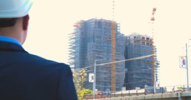 Genç bir adam (erkek) mimar, takım elbiseli bir işadamı, elinde tabletli bir klasör (cep telefonu) olan bir ceket içinde inşaat alanını ve iş binasını çeşitli eylemlerle kontrol ediyor..