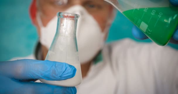 炼金术士 手持烧瓶 测试试管的不同成分 在白色大衣 呼吸器 护目镜 橡胶白色手套 绿色背景 — 图库视频影像