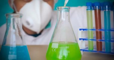 Simyacı, şişeler tutan, bir testin farklı kompozisyonlar, beyaz önlük, solunum, Gözlük, beyaz eldivenleri, yeşil bir arka plan ile test tüpleri testleri.