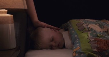 Küçük bir çocuk (oğlu) tatlı, sarışın uyuyor, anne karanlıkta, oğlunu öper. Konsept: çocuklar, çocuklar, bebek.