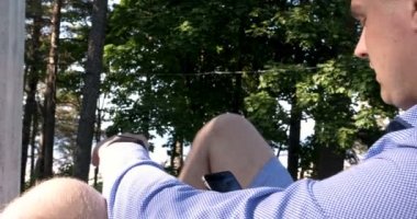 Adam parkta bir ağacın altında, suyun yanında bir tablet bilgisayar kullanıyor. Arka planda şehir manzarası. Seçici odak, tonda görüntü, yakışıklı genç beyaz turist adam mutlu ve bir selfie alarak heyecanlı, çekici genç Kafkas turist öğrenci adam hav