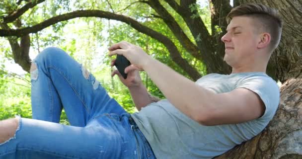 在公园的一棵树下 在水边 男子使用平板电脑 背景上的城市景观 选择性的焦点 调子的形象 英俊的年轻高加索旅游男子高兴和兴奋地采取自拍 有吸引力的年轻高加索旅游学生男子哈夫 — 图库视频影像
