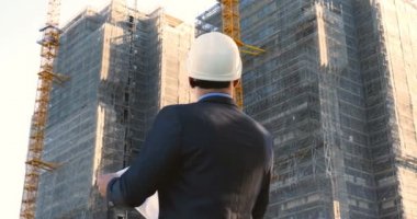 Başarılı genç mühendis, mimar, müteahhit, iş adamı portresi, beyaz bir kask takıyor, takım elbise giyiyor, elinde not defteri, gökdelen arka planı ve bina var..