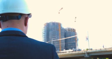 Genç bir adam (erkek) mimar, takım elbiseli bir işadamı, elinde tabletli bir klasör (cep telefonu) olan bir ceket içinde inşaat alanını ve iş binasını çeşitli eylemlerle kontrol ediyor..